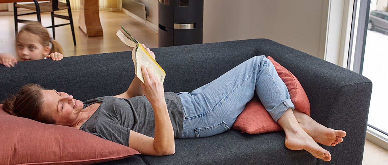En dame ligger i en sofa og leser en bok, og et barn er i bakgrunnen og titter over sofaen